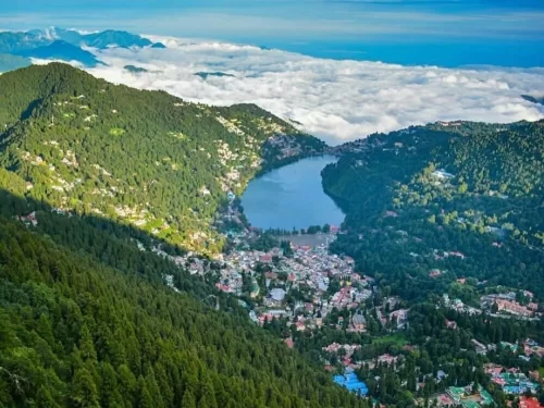 Aerial panoramic of Naini Lake Nainital during cloudy sunny day, featuring pear-shaped turquoise waters amidst sea of clouds and forested hills, perfect romantic experience Uttarakhand tour package.