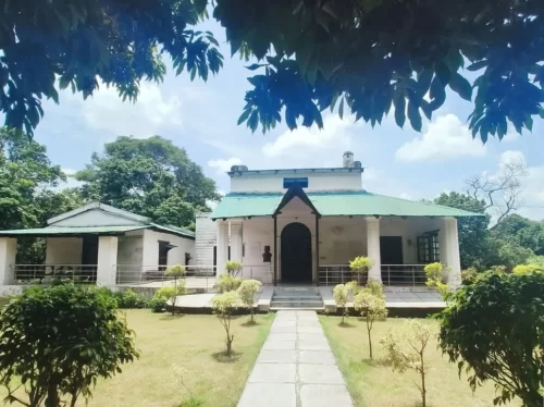 Jim Corbett Museum heritage bungalow green tin roof white walls, colonial architecture arched entrance balcony potted plants lawn trees cloudy sky, perfect Uttarakhand tour packages.