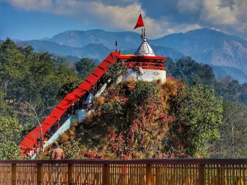 Garjiya Devi Temple Ramnagar hilltop closeup, featuring white temple red flag ramp saffron marigold decorations bamboo fence forested mountains cloudy skies, perfect Uttarakhand tour packages.