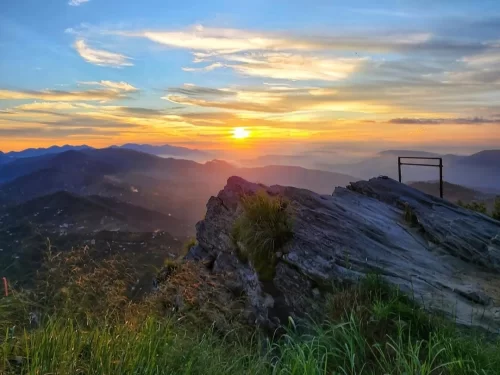 Chauli ki Jali at sunset in Mukteshwar during golden hour, featuring rocky ledge metal railing, orange sun hills valleys, perfect dramatic vista, Uttarakhand tour packages.