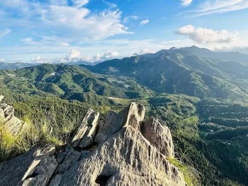 Chauli ki Jali panoramic viewpoint in Mukteshwar during sunny weather, featuring rocky outcrop, green hills Himalayan peaks clouds, perfect scenic trek, Uttarakhand tour packages.