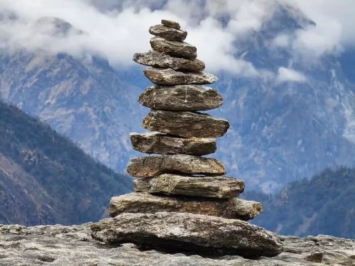 Khaliya Top Munsiyari during misty weather, featuring stone cairn stack, Himalayan peaks, clouds, perfect trekking adventure, Uttarakhand tour packages.