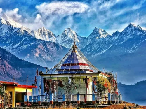 Nanda Devi Temple Munsiyari during sunny day, featuring colorful pagoda roof temple prayer flags railing small buildings foreground snow-capped Himalayan peaks brown hills clouds blue sky, perfect spiritual trekking experience Uttarakhand tour packages.