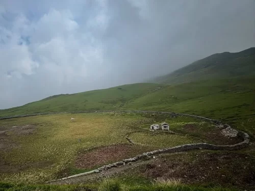 Stone huts at Gurso Bugyal during misty cloudy weather, featuring green alpine meadows stone walls hills, perfect adventure experience Uttarakhand tour packages.