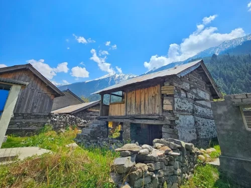 Bagori Village wooden cottages during sunny day Uttarkashi, featuring rustic stone wood houses grass stone walls snowy distant mountains blue sky, perfect Uttarakhand tour packages.