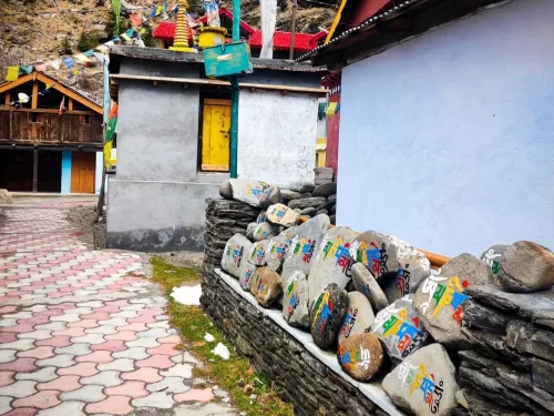 Bagori Village prayer stones during cloudy winter day Uttarkashi, featuring colorful mani stones wall, traditional houses prayer flags pink cobbled path snowy backdrop, perfect Uttarakhand tour packages.