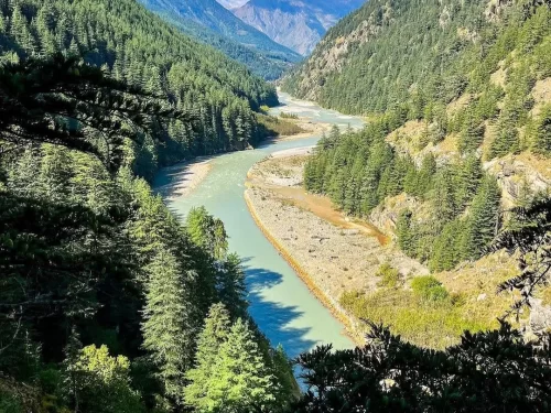 Harshil Valley panoramic during sunny day, featuring turquoise river winding through pine forests, rocky mountains, perfect adventure experience Uttarakhand tour packages. 