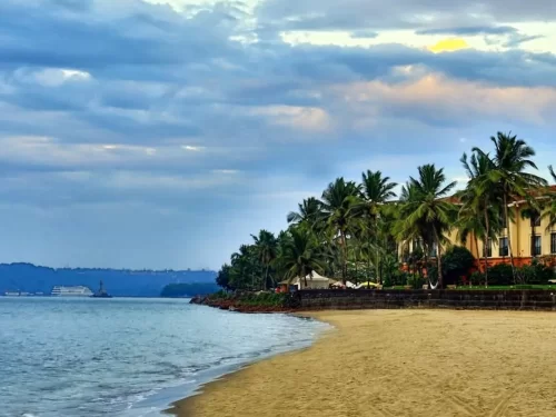 Sandy Miramar Beach Goa at golden hour with palm-fringed luxury resort Mandovi River cruise ship backdrop cloudy sunset sky gentle waves, perfect beach experience Goa beach resort stay package.
