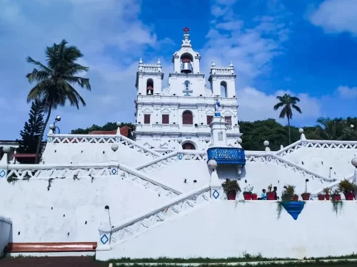 Bright Our Lady of Immaculate Conception Church Panjim Goa under partly cloudy blue sky with visitors potted plants on sweeping white staircase palm trees blue balustrade lamps greenery accents, perfect heritage experience Panjim Goa church feast Immacula