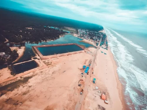 Chandrabhaga Beach Odisha aerial view featuring blue water tank sandy shore road palm trees fishing boats cloudy sky near Konark Sun Temple, perfect beach experience for Konark Chandrabhaga tour package.