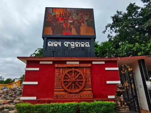 Odisha State Museum Bhubaneswar grand red entrance gate Odisha script LED tribal dancers chariot wheel cloudy sky, perfect Odisha heritage culture tour package. 