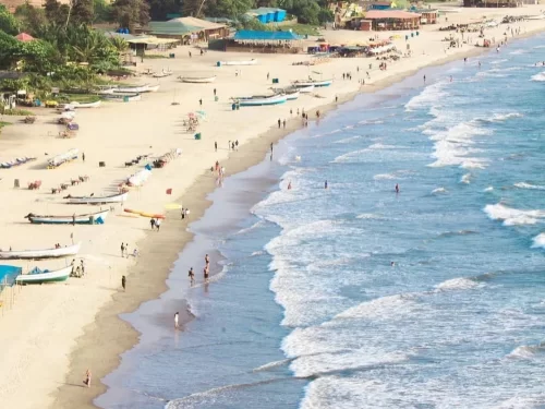 Crowded beach scene at Arambol Beach Goa during sunny day, featuring fishing boats, beach shacks, palm trees, gentle waves, perfect hippie experience with Arambol Goa tour package.