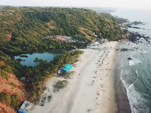 Aerial view of Arambol Beach Goa during misty morning, featuring Sweet Water Lake, green hills, beach shacks, crowds, waves, perfect bohemian experience with Arambol tour package. 