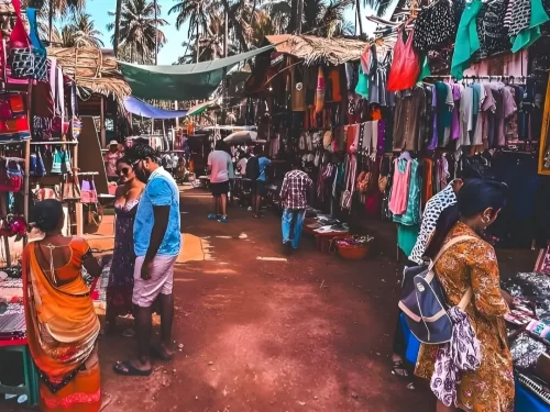 Shoppers browsing colorful clothes at Anjuna Flea Market in Goa during sunny day, featuring thatched stalls palm trees, perfect shopping adventure experience with Goa beach tour package.
