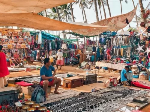 Vibrant stalls with clothes jewelry at Anjuna Flea Market in Goa during sunny day, featuring thatched roofs palms red earth, perfect shopping adventure experience with Anjuna flea market tour package