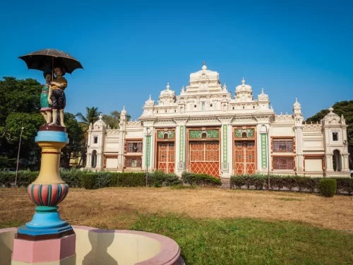 Jaganmohan Palace in Mysore during sunny daytime, featuring ornate facade and umbrella statue fountain, perfect heritage Karnataka tour package
