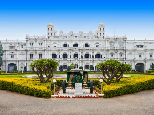 Jai Vilas Palace Gwalior during clear afternoon, featuring white Italianate facade with domed towers before manicured gardens showcasing silver royal carriage display amid yellow flower borders, perfect heritage experience in Madhya Pradesh tour package.