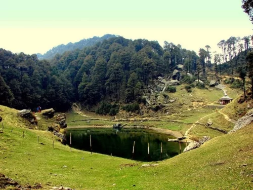 Scenic view of Jalori Pass featuring a serene mountain lake surrounded by grassy slopes, dense pine forests, rocky outcrops, and a small temple along a walking trail in the Himalayas.