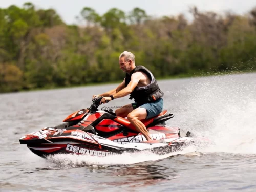 Jet Skiing man riding a red jet ski at high speed across a lake wearing a life jacket with water splashing around him.