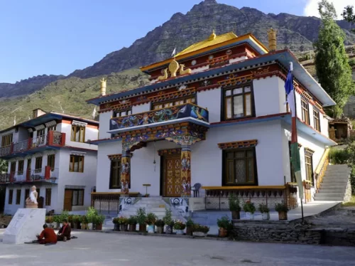 Front view of Kardang Monastery in Keylong, featuring a colorful Tibetan-style building with ornate entrance, golden roof details, prayer flags, potted plants, and mountains rising in the background.