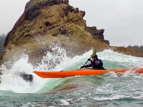 Kayaking person paddling an orange kayak through powerful ocean waves near rocky cliffs with water splashing around.
