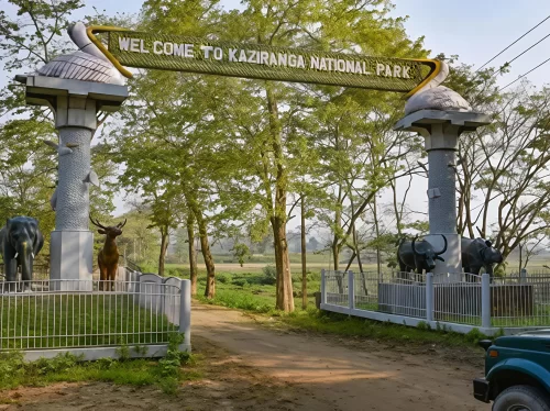 Kaziranga National Park Assam India main entrance gate golden yellow welcome sign white stork statues tall pillars elephant deer statues green trees paddy fields jeep parked dirt road iconic wildlife park gateway, perfect Northeast India tour packages.