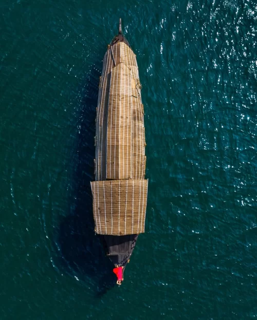 Top aerial view of a traditional Kerala houseboat cruising through deep blue waters, with its woven roof structure clearly visible from above.