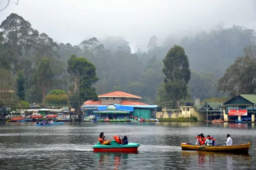Kodai Lake in Kodaikanal during misty day, featuring colorful pedal and row boats with tourists, lakeside boat house and pine-covered hills, perfect family Tamil Nadu tour package