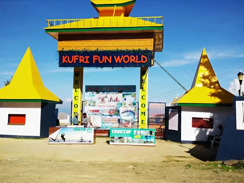 Entrance gate of Kufri Fun World featuring a colorful yellow and green structure with bold signage, ticket counters in front, and clear blue sky in the background.