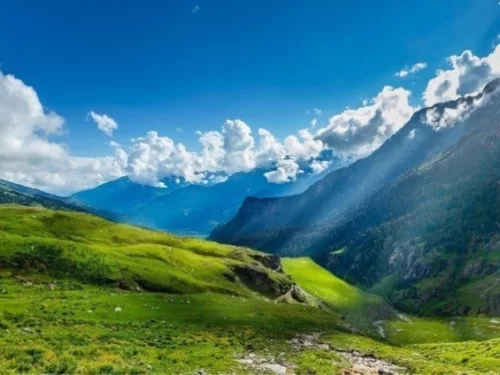 Scenic Kullu Valley featuring lush green meadows, rolling hills, and sun rays over the Himalayan mountains under a blue sky.