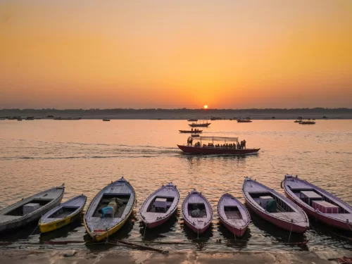 Boats at Kusum Sarovar during sunset, featuring colorful rowboats, distant passenger boat, golden sun rays, perfect romantic experience with Mathura Vrindavan tour package. 
