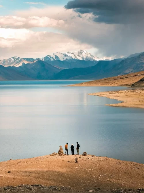 Travelers standing at Tso Moriri Lake in Ladakh with snow-capped Himalayan peaks and calm blue waters, a breathtaking view in Ladakh tour packages