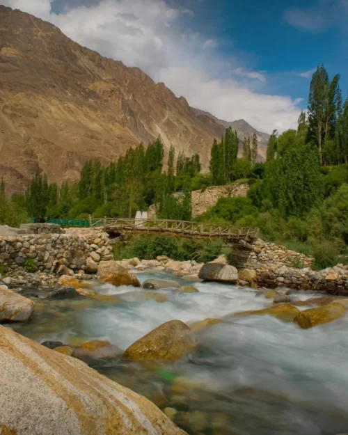 Scenic Turtuk village in Ladakh with flowing river, stone bridge and lush greenery against rugged mountains, a hidden gem in Ladakh tour packages