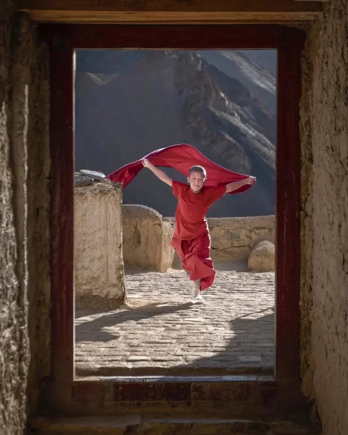 Buddhist monk in red robes walking through Lamayuru Monastery doorway with Himalayan mountains in Ladakh tour packages