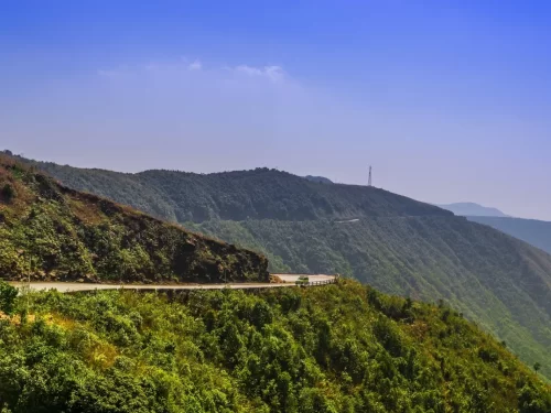 Laitlum Canyons Shillong Meghalaya winding mountain road hairpin bends lush green hills blue sky tower Shillong Peak background scenic drive viewpoint near canyons, perfect Meghalaya tour packages.