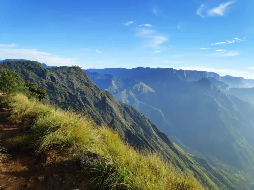 Laitlum Canyons Shillong Meghalaya scenic ridgeline trail lush grassy slopes dramatic layered mountain ridges deep valleys blue sky scattered clouds golden sunlight misty horizons trekking viewpoint, perfect Meghalaya tour packages.