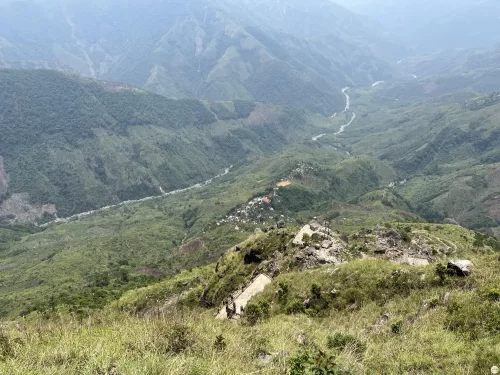 Laitlum Canyons Shillong Meghalaya panoramic valley view lush green hills winding river villages terraced fields distant blue mountains cloudy sky foreground grass rocky outcrop tiny figures viewpoint, perfect Meghalaya tour packages.