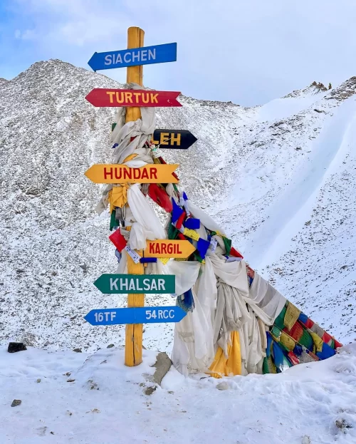 Signpost at Khardung La Top in Ladakh pointing toward Siachen, Turtuk and Leh amid snow-covered mountains, an iconic stop in Ladakh tour packages