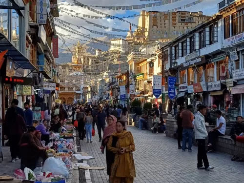 Sunny Leh Market street lined with shops, prayer flags overhead and Leh Palace in distant backdrop with diverse crowds, perfect India tour package. 