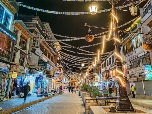 Nighttime Leh Market illuminated by street lamps, fairy lights and hanging baskets under prayer flags with sparse evening crowds, perfect India tour package. 