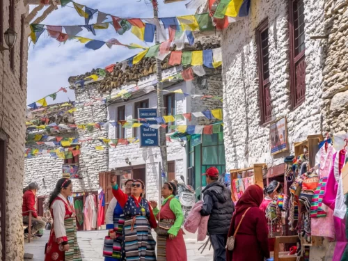 Lively Leh Market lane with prayer flags overhead, locals in traditional attire shopping vibrant textiles amid white-washed Ladakhi buildings, perfect India tour package.
