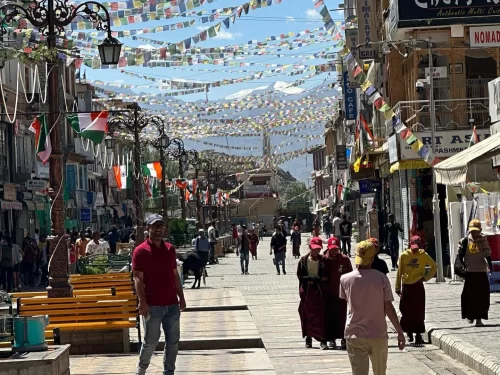Bustling Leh Market street under prayer flags and international banners with diverse crowds, monks and shops, perfect India tour package in Ladakh. 