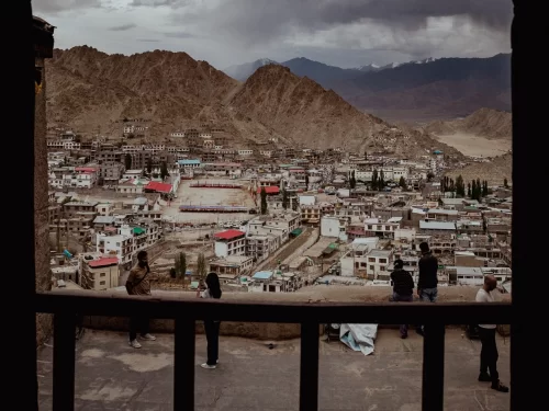 Leh Palace Ladakh panoramic cityscape view from palace balcony showcasing traditional Ladakhi buildings nestled between rugged mountains under cloudy skies, tourists observing, ultimate Himalayan cultural tour package. 