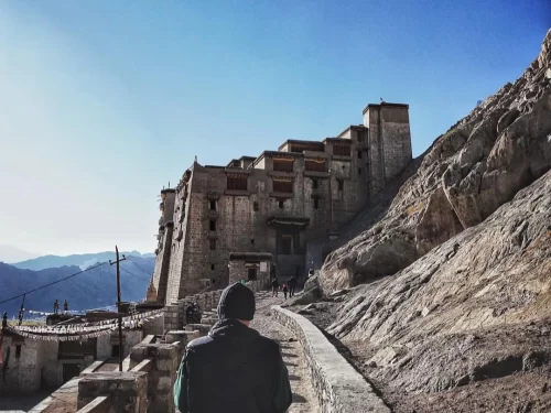 Leh Palace Ladakh dramatic multi-tiered white stone fortress towering over rocky terrain path with silhouetted hiker visitors distant snowy mountains blue sky, epic Himalayan palace trek tour package.