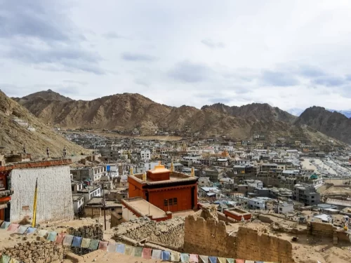 Leh Palace Ladakh elevated panoramic view over traditional Ladakhi town with colorful prayer flags red roofed buildings ruins against dramatic rugged mountains cloudy skies, immersive Himalayan cultural adventure tour package.