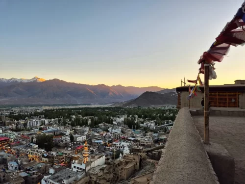 Leh Palace terrace view over Leh town with colorful buildings along Indus Valley, snow-capped Stok Kangri mountains at sunset, fluttering prayer flags in foreground, perfect India tour package.