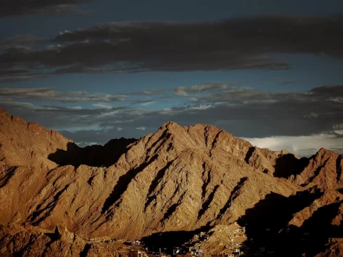 Leh Palace stunning sunset vista of rugged brown Ladakh mountains silhouetted against dramatic dark clouds and blue twilight sky, perfect India tour package.