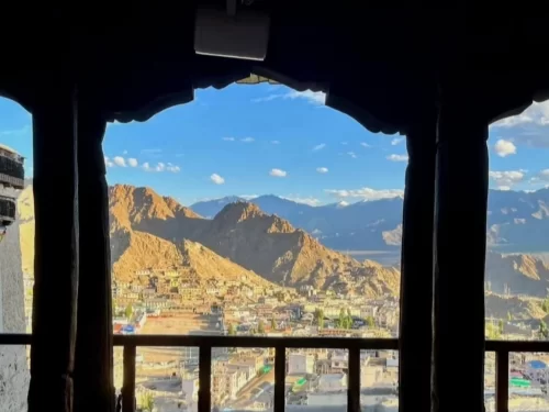 Leh Palace wooden framed balcony panoramic view of Leh town nestled in arid valley with distant rugged mountains under partly cloudy blue sky, perfect India tour package.