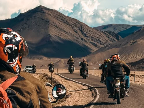 Leh City Road Ladakh dynamic biker group convoy roaring along dusty highway amidst towering dramatic brown mountains cloudy blue skies rugged adventure rider POV, exhilarating Himalayan motorcycle safari biking expedition tour package