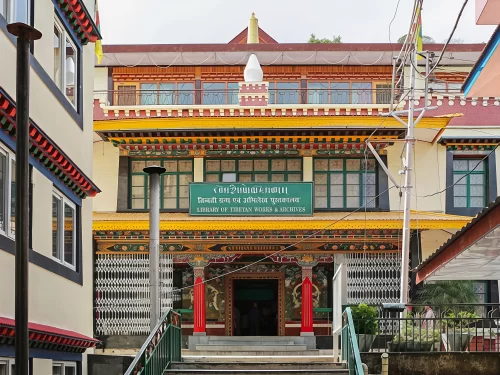 Library of Tibetan Works and Archives in Dharamshala featuring a traditional Tibetan-style building with colorful wooden carvings, green signboard at the entrance, and decorative pillars leading up a short staircase.